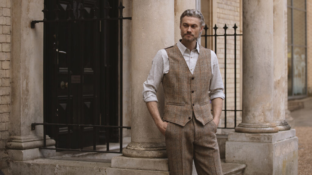 Man leaning against a pillar outside a stately home, wearing a tweed waistcoat and trousers and checked shirt from Walker & Hawkes.