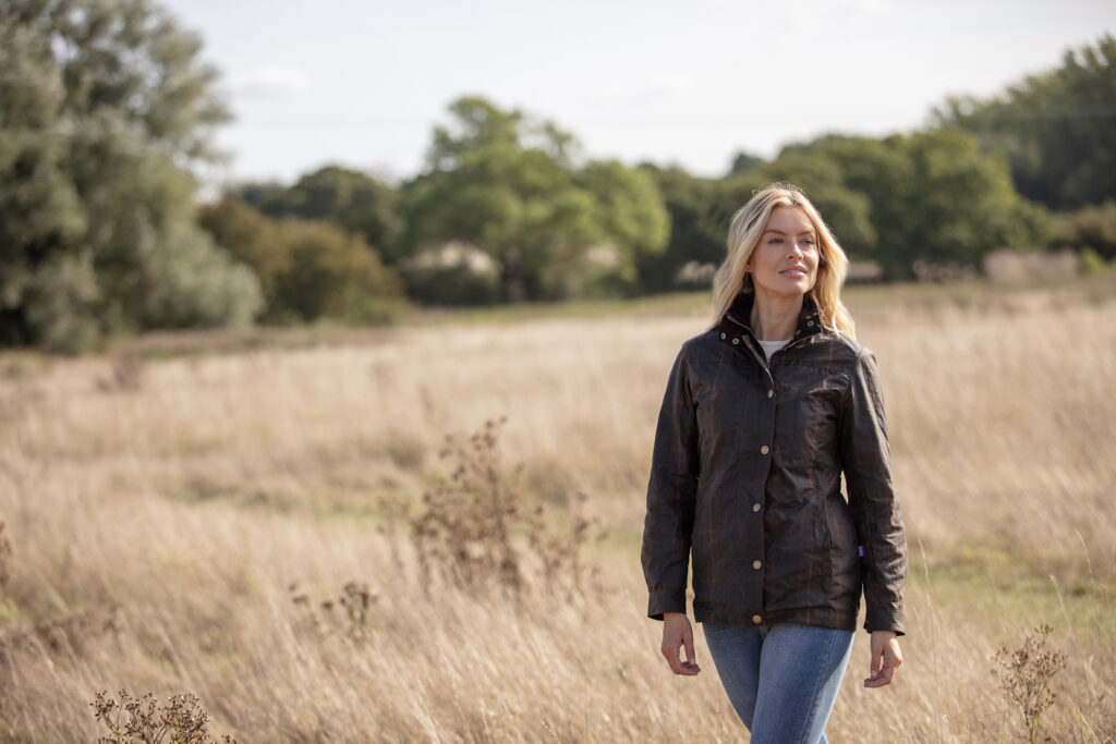 Woman walking through dried long grass, on a breezy day, wearing a Walker & Hawkes wax jacket.